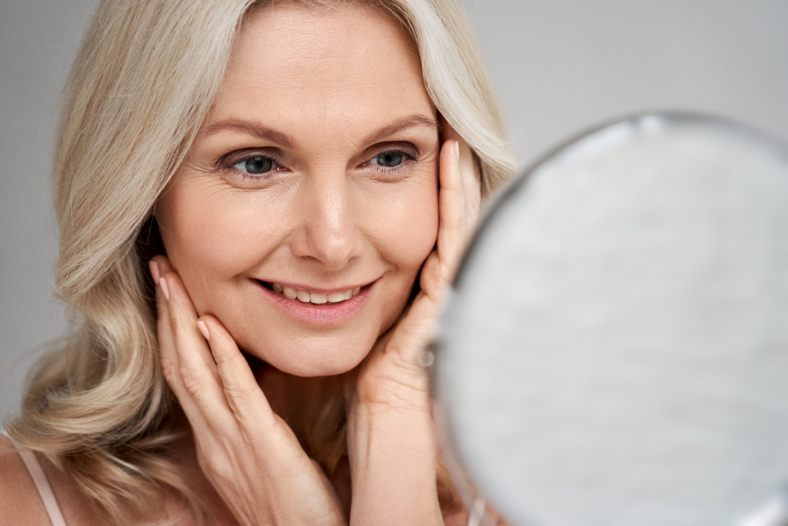 Smiling mature woman admiring her smooth complexion in the mirror after using medical-grade skincare